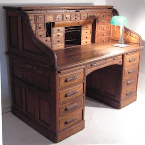May include: A large, antique wooden roll-top desk with numerous drawers and compartments. The desk is made of dark-stained wood and features a curved roll-top design. A small green glass lamp sits on the desk surface. The desk has a classic, vintage aesthetic.