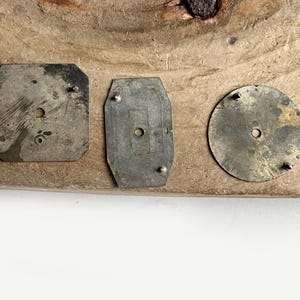 May include: Three weathered metal components with a tarnished finish. The set includes a square plate with clipped corners, a rectangular plate with rounded edges, and a circular disc. Each piece features small protruding pins and a center hole.