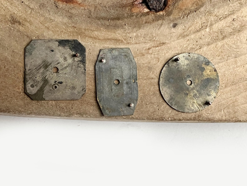May include: Three weathered metal components with a tarnished finish. The set includes a square plate with clipped corners, a rectangular plate with rounded edges, and a circular disc. Each piece features small protruding pins and a center hole.