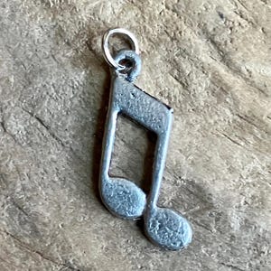 May include: A small, silver-colored charm in the shape of a musical eighth note. The charm has a loop at the top for attaching to a necklace or bracelet. The charm is made of a metal with a slightly textured surface.