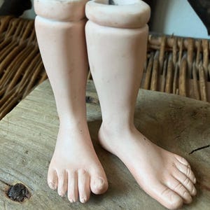 May include: A pair of pale, flesh-toned doll legs made of smooth material. The legs are positioned upright, showing detailed toes and rounded calves. Each leg features a hollow, cylindrical opening at the top, designed for attachment to a doll body.