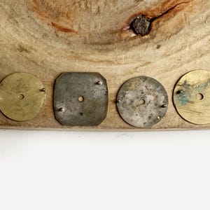 May include: Four small metal components arranged in a row. Two are circular brass discs, one is a square-shaped silver-toned piece with rounded corners, and one is a circular silver-toned disc. Each piece features a central hole and small metal pins.