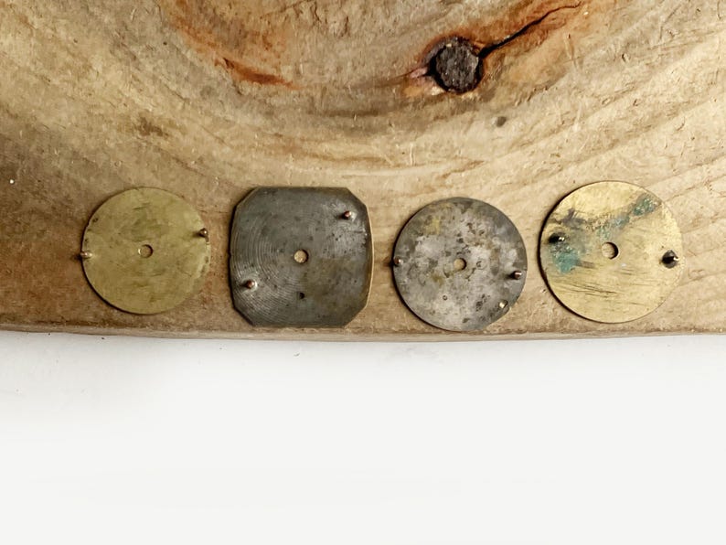May include: Four small metal components arranged in a row. Two are circular brass discs, one is a square-shaped silver-toned piece with rounded corners, and one is a circular silver-toned disc. Each piece features a central hole and small metal pins.