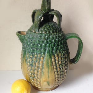 May include: A green ceramic pitcher with a textured, pineapple-like design. The pitcher has a handle and a lid with a decorative top. A yellow lemon sits next to the pitcher.