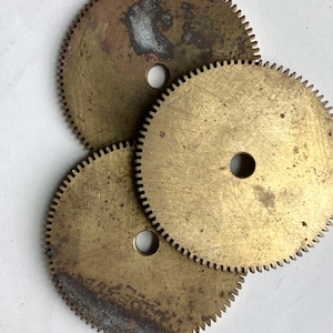 May include: Three antique brass gears with toothed edges and central holes. The gears have a worn, aged appearance with areas of discoloration and patina, suggesting they are vintage clock or machinery parts.