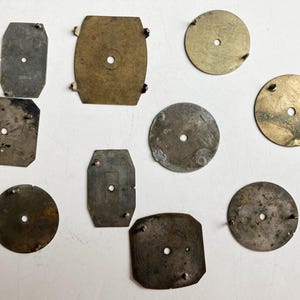May include: A collection of ten assorted metal watch dial plates in various shapes including circular, rectangular, and square forms. Each plate features a central hole and small protruding metal pins on the surface, showing signs of age and wear.
