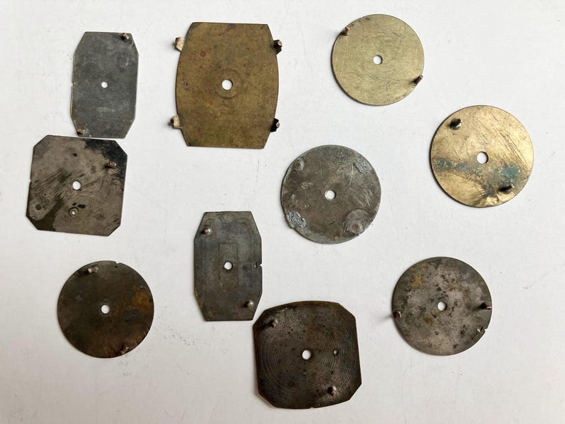 May include: A collection of ten assorted metal watch dial plates in various shapes including circular, rectangular, and square forms. Each plate features a central hole and small protruding metal pins on the surface, showing signs of age and wear.