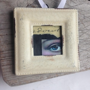 May include: A square, cream-colored frame with a distressed finish and a white ribbon hanger. The center features a collage with a blue eye, dark hair, and the handwritten word advent in black ink on a yellowed paper background.