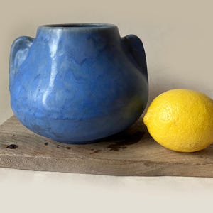 May include: A blue ceramic vase with two handles sits on a wooden plank next to a yellow lemon. The vase has a rounded shape and a textured surface. The lemon is smooth and bright yellow.