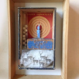 May include: A shadow box display featuring a vintage Daisy Targeteer box with a red target, a light blue figurine, a clear tube of white spheres, and a blue wooden block. Below the block is a white fabric piece printed with black deer silhouettes.