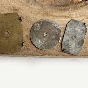 May include: Three metal components for watch repair or assembly. One rectangular brass piece with four small prongs, one circular silver-toned disc with a central hole, and one rectangular silver-toned plate with two small prongs and a central hole.