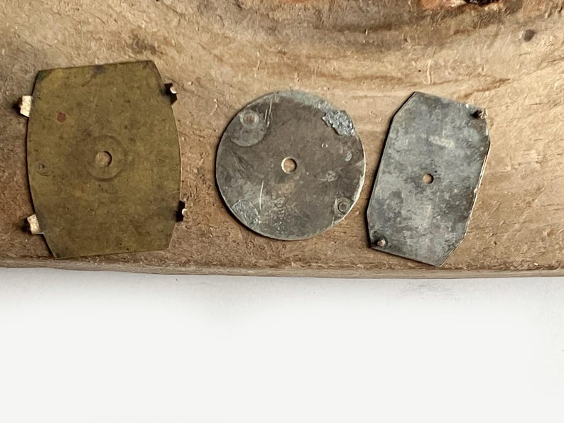 May include: Three metal components for watch repair or assembly. One rectangular brass piece with four small prongs, one circular silver-toned disc with a central hole, and one rectangular silver-toned plate with two small prongs and a central hole.