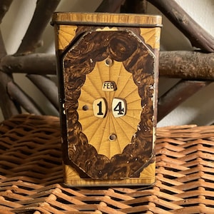 May include: Vintage metal perpetual calendar with a decorative wooden design. The calendar displays "FEB 14" on its face. The calendar is rectangular and sits on a woven surface.