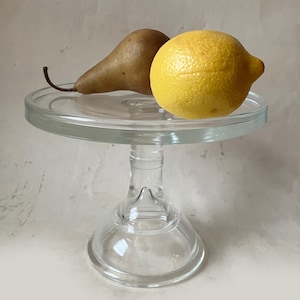 May include: A clear glass pedestal cake stand with a round flat top and a sturdy base. Resting on the surface of the stand are a single brown pear with a curved stem and a bright yellow lemon, both positioned side by side on the glass plate.