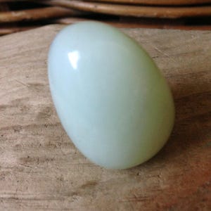 May include: A smooth, oval-shaped, pale green stone. The stone has a glossy finish and reflects light. The object is resting on a wooden surface with visible grain. The overall aesthetic is natural and minimalist.