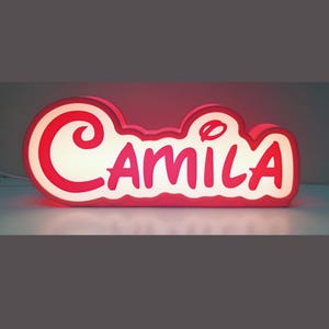 Può includere: Un cartello illuminato rosso e bianco con il nome "Camila" in un carattere fantasioso.