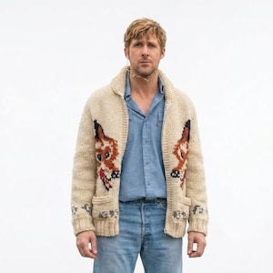 May include: A cream-coloured knitted cardigan with a shawl collar and two front pockets. The sweater features a cartoon fox design in shades of brown, black, and red on the front. The person is wearing a blue button-down shirt and blue jeans.
