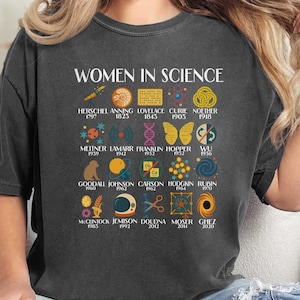 Puede incluir: Camiseta gris oscuro con el texto "WOMEN IN SCIENCE" e ilustraciones de científicas y sus descubrimientos. Presenta nombres como Curie, Franklin y Goodall, con los años asociados. Una camiseta gráfica única y educativa.