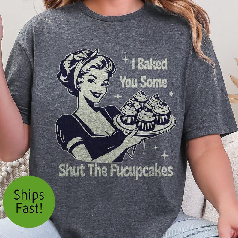 以下が含まれることがあります： レトロなイラストが特徴の濃いグレーのTシャツ。女性がカップケーキのプレートを持っています。Tシャツのテキストは「I Baked You Some Shut The Fucupcakes」と書かれています。デザインは白黒です。