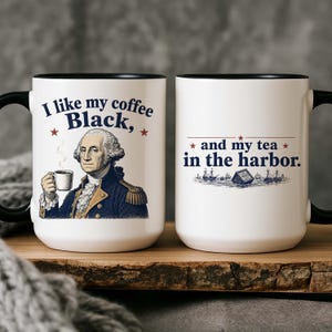 May include: A white ceramic mug with a black handle and rim, featuring an illustration of George Washington holding a coffee cup and the text I like my coffee Black, and my tea in the harbor, with a graphic of ships and a crate in the water.