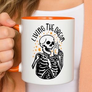 Op de afbeelding: Witte keramische mok met een oranje binnenkant en handvat. De mok heeft een zwart-wit skelet illustratie met een koffiekopje, omringd door sterren, met de tekst "LIVING THE DREAM".