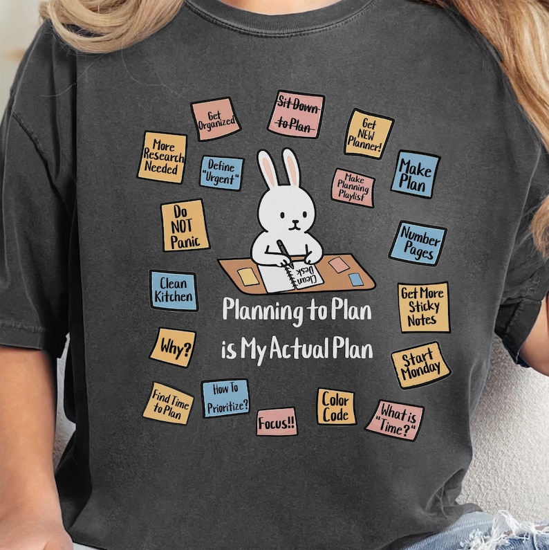 May include: Dark gray t-shirt with a graphic of a bunny at a desk, surrounded by sticky notes with planning-related phrases. The text on the shirt reads "Planning to Plan is My Actual Plan."