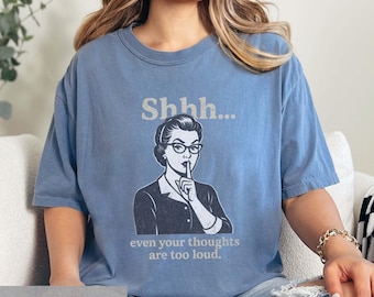 Op de afbeelding: Blauw spijkerbroekkleurig T-shirt met de tekst "Shhh..." boven een afbeelding van een vrouw met een bril, vinger op haar lippen. Onder de afbeelding staat de tekst "even your thoughts are too loud."