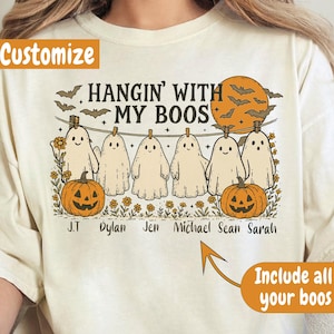 Pode incluir: Camiseta creme com um gráfico temático de Halloween. O design apresenta fantasmas de desenhos animados pendurados em uma corda, abóboras, morcegos e o texto "HANGIN' WITH MY BOOS". Os nomes estão listados abaixo. A camiseta é personalizável.