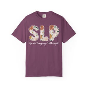 May include: A plum-colored short-sleeve t-shirt featuring the large letters SLP filled with a floral pattern of white, orange, and green flowers. Below the letters, the text Speech Language Pathologist is printed in a white cursive script font.