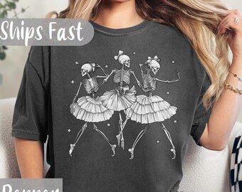 Può includere: T-shirt grigio scuro con un'illustrazione in bianco e nero di tre ballerine scheletriche con tutù e fiocchi. Il testo "Pepper" è in basso sulla maglietta. La maglietta ha un aspetto vintage e usurato.