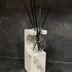 Peut inclure: Un diffuseur de parfum à bâtonnets en céramique blanche avec une découpe incurvée et des accents dorés mouchetés. Un flacon sphérique en verre transparent est placé dans le support, contenant plusieurs bâtonnets noirs fins vers le haut.