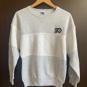 May include: A gray and white sweatshirt with a Russell Athletic logo and a Philadelphia Flyers logo on the front. The sweatshirt has a white band across the chest.
