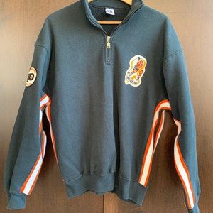May include: A dark green 1/4 zip sweatshirt with orange and white stripes on the sleeves. The sweatshirt has a large embroidered logo on the front with a hockey player and the text "Philadelphia Flyers".
