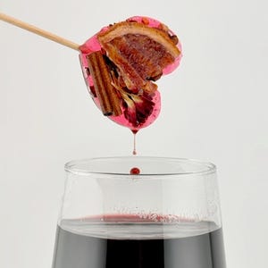 May include: A wine glass filled with dark red liquid, with a heart-shaped garnish on a wooden stick above it. The garnish includes a slice of orange, cinnamon, and a pink coating. A drop of liquid falls from the garnish.