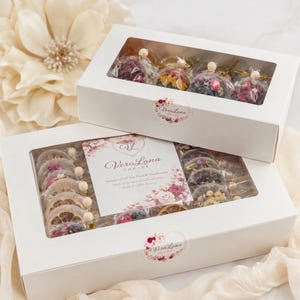 May include: Two white rectangular boxes with clear windows containing individually wrapped tea spheres filled with dried flowers and fruit slices. The labels feature the text VeraLana TREATS and floral graphics. The boxes are arranged together.