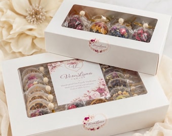 May include: Two white rectangular boxes with clear windows containing individually wrapped tea spheres filled with dried flowers and fruit slices. The labels feature the text VeraLana TREATS and floral graphics. The boxes are arranged together.