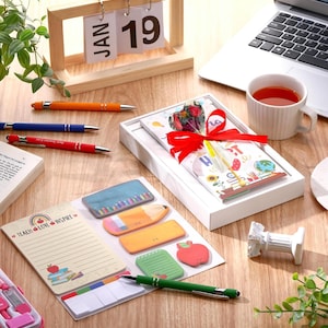 Peut inclure: Un calendrier de bureau en bois affiche "JAN 19". Le bureau contient également des stylos orange, bleu et rouge, ainsi qu'un coffret cadeau noué d'un ruban rouge. Il y a aussi des post-it, une tasse de thé et un ordinateur portable.
