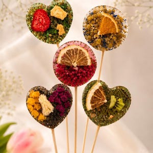 May include: Five lollipops with fruit inclusions, some heart-shaped, others round, on wooden sticks. The lollipops contain dried fruit pieces, including strawberries, oranges, and kiwi. The lollipops are presented on a white surface.