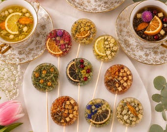 May include: A collection of eight round, clear lollipops embedded with dried flowers, herbs, and citrus slices, each on a thin wooden stick. The lollipops are arranged on a white board alongside three floral teacups filled with tea and citrus.