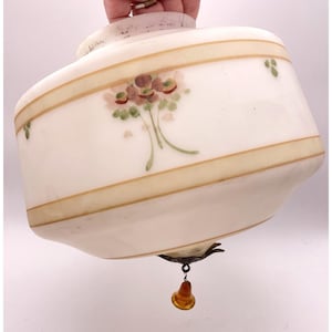 May include: An antique, off-white glass lampshade with a rounded rectangular shape. It features a painted floral design in shades of pink, green, and brown, with a gold-colored band. A small, amber-colored glass pendant hangs from the bottom.