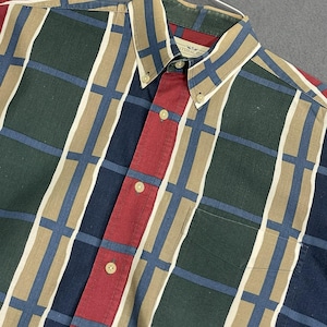 May include: Button-down shirt with a plaid pattern in shades of green, blue, tan, and red. The shirt features a button-down collar, a chest pocket, and a red vertical stripe down the center.