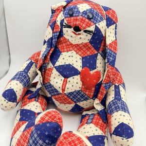 May include: A stuffed rabbit toy crafted from fabric with a patchwork pattern of red, white, and blue geometric shapes. The rabbit features two dark blue button eyes, black embroidered whiskers, and a small red fabric heart sewn onto its chest.