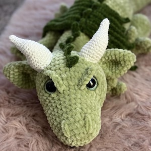 Könnte beinhalten: Ein gehäkeltes Drachen-Stofftier aus weichem hellgrünem Garn mit zwei markanten weißen Hörnern, dunkelgrünen strukturierten Flügeln und großen schwarzen Augen mit einem dezenten Glitzereffekt. Das Wesen hat einen langen Schwanz und eine genähte Schnauze.