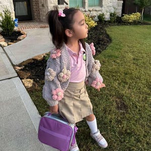 May include: A young person wearing a gray cardigan adorned with large, pink and cream-colored crocheted flowers. The outfit includes a pink polo shirt, khaki skirt, and a purple lunch bag. The person is standing on a grassy area.