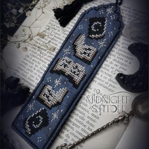 Puede incluir: Un marcapáginas artesanal con fondo azul oscuro, con símbolos bordados en negro y beige. El marcapáginas tiene una borla negra y está sobre un libro abierto. El texto "MIDNIGHT STITCH" es visible.