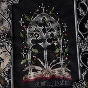 Peut inclure: Broderie au point de croix dans un cadre argenté décoratif. Le tissu noir présente un motif de livre ouvert avec une arche gothique et des éléments floraux blancs, verts et oranges. Le cadre a un motif floral.
