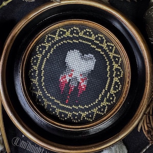 Puede incluir: Una obra de arte en punto de cruz enmarcada con un diseño de diente con detalles rojos sobre un fondo negro. La obra está enmarcada en un marco redondo negro y dorado. La imagen incluye el texto "@midnight.xstitch".