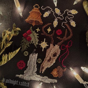 May include: Cross-stitch design on black fabric featuring a bell, berries, and the word "Good" in a stylized font. The design is surrounded by string lights and a brass bell. The text "@midnight.xstitch" is visible.