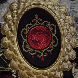 May include: An oval, framed cross-stitch artwork featuring a red moon design with gold accents on a black background. The frame is gold and has a scalloped edge. The artwork is displayed on a dark surface.