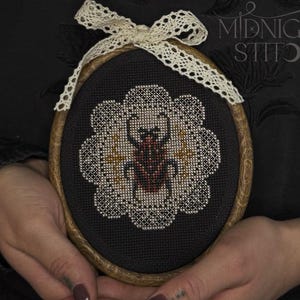 Può includere: Ricamo a punto croce ovale con un disegno di scarabeo rosso e marrone, circondato da un motivo floreale bianco su sfondo nero. È incorniciato in un cerchio di legno e rifinito con un fiocco di pizzo color crema. Il testo "MIDNIGHT STITCH" è visibile.